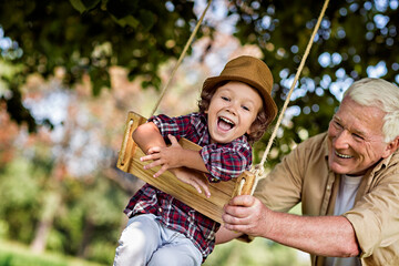 Young boy and his grandfather spending time in the park on a swing - obrazy, fototapety, plakaty