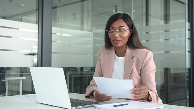 Professional young African American business woman hr manager team leader executive wearing suit working in office looking at laptop, holding paper having video call hybrid virtual online meeting.