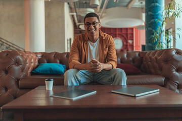 Smiling male freelancer sitting on sofa and looking at camera during working day in coworking