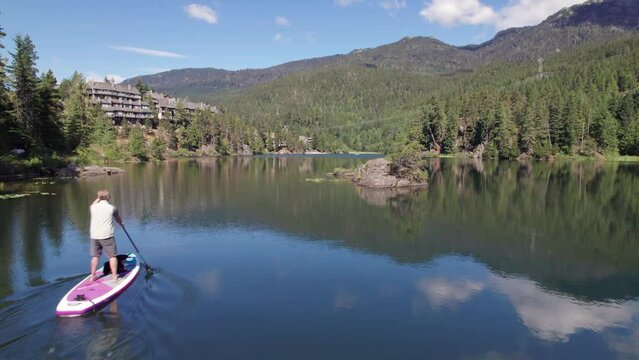 Man paddle boarding on a pristine mountain lake on sunny day with his dog, stand up paddle, mountains, Whistler, BC, water sport. 4K 24FPS PRORES 422