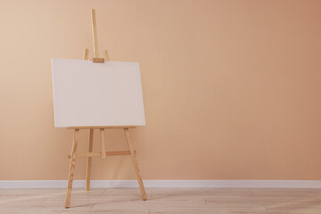 Wooden easel with blank canvas near beige wall indoors. Space for text