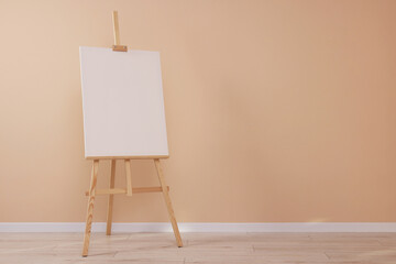 Wooden easel with blank canvas near beige wall indoors. Space for text