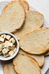 Top view of marinated feta cheese cubes and slices of toasted bread on a white plate. Cheese cubes in a ramekin with bread, flat lay of feta cheese cubes and slices of toast