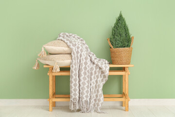 Fototapeta na wymiar Wooden bench with blanket, cushions and plant in wicker basket near green wall