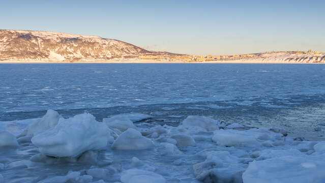 Winter seascape. Icy shore and ice-covered sea bay. Freezing water. Port city in the distance. Cold frosty winter weather. Nagaev Bay, Sea of Okhotsk. Magadan, Magadan Region, Far East of Russia. - Powered by Adobe