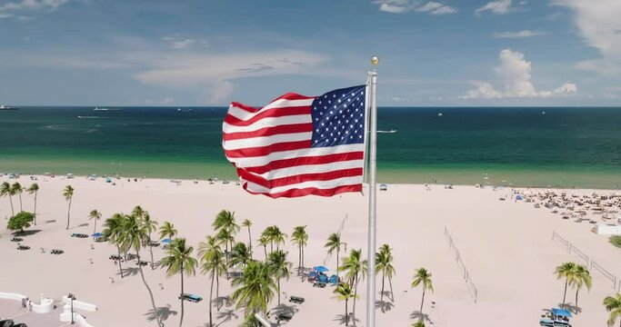 Aerial orbital shot of American flag against palm trees on the beach and ocean background. Slow motion