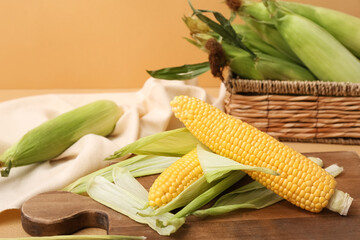 Wooden board and wicker basket with fresh corn cobs on brown background