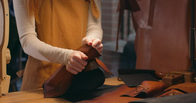 young woman in apron standing by table, touching pieces of leather, bending it, studying and comparing materials. Slow motion