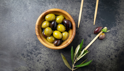 Fresh olives in wooden bowl on dark stone table. Black and green olive with pickers or sticks from top view.