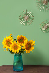 Vase with beautiful sunflowers on table and decor hanging on green wall in room