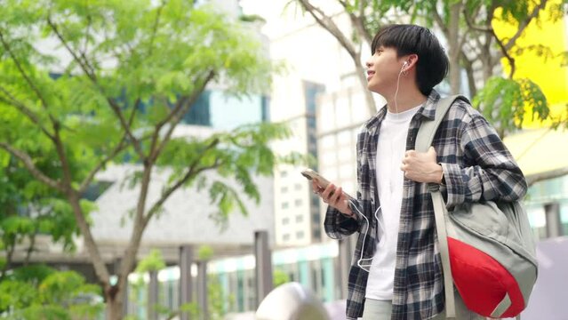 4K Young Asian man university college student walking in the city with listening to the music on earphones and using mobile phone app chatting online text message or social media with online network.