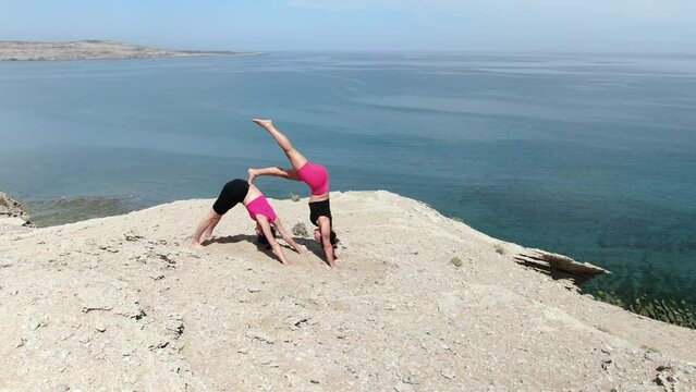 Yoga in nature, two people practicing yoga posture on the edge of a cliff, sorrounded by a beautiful landscape, ocean background. Aerial orbital shot.