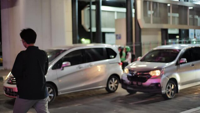 male pedestrian waits to cross under Jakarta's ASEAN pedestrian bridge at night. There are cars and motorcycles stopped in front of him. The entire bridge is decorated with twinkling lights. Medium s