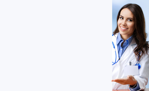 Portrait image of smiling female doctor showing blank signboard with copy space area for some text, at office. Young brunette woman in medical, clinic, healthcare concept