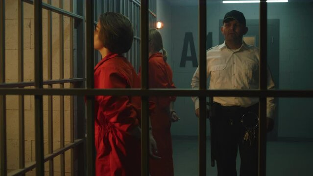 Two female prisoners, inmates in orange uniforms stand facing the metal bars in front of prison cells. Prison officer walks, watches women criminals in jail. Detention center or correctional facility.