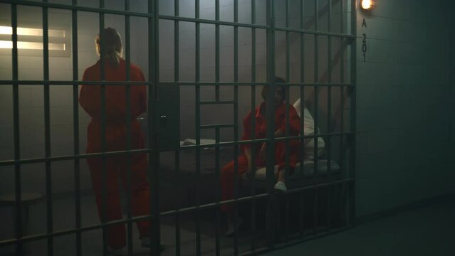 Two female prisoners in orange uniforms talk. One woman sits on the bed, another walks in prison cell. Women serve imprisonment terms for crimes in jail, detention center or correctional facility.