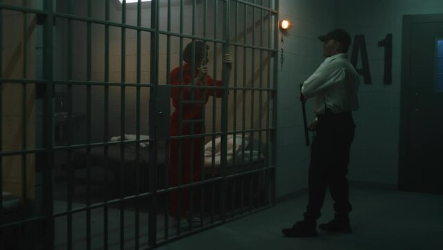 Prison employee with police baton talks to female prisoner in orange uniform in prison cell. Women criminals serve imprisonment terms for crimes in jail. Detention center or correctional facility.