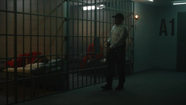 Prison officer with police baton talks to female prisoners in orange uniforms through metal bars. Women serve imprisonment terms in prison cells. Jail, detention center or correctional facility.