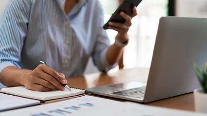 Close up of businesswoman is searching data on smartphone while working on laptop and writing notes