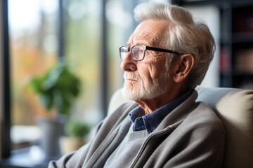 Fototapeta premium old man sitting in an armchair and looking through the window Lonely old man sitting at home near window pandemic. Thoughtful retiree man abandoned at the nursing home.