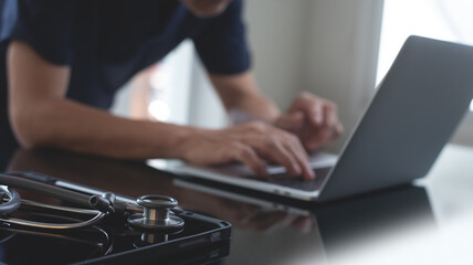 Close up of male doctor working and typing on laptop computer at doctor's office with stethoscope and digital tablet on table, doctor staff online meeting via laptop