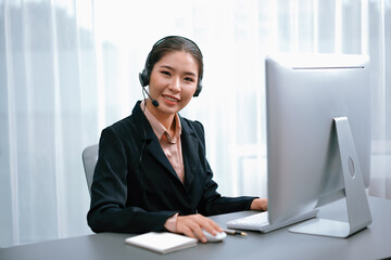 Asian call center with headset and microphone working on her workspace looking at camera. Female...