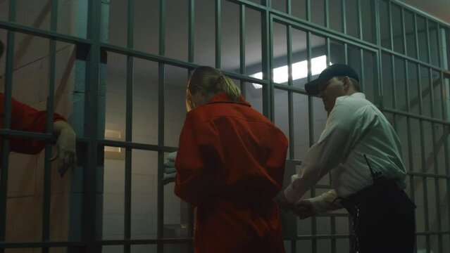 Prison employee leads female prisoner in orange uniform to prison cell, locks inside, puts off handcuffs. Women serve imprisonment terms for crimes in jail. Detention center or correctional facility.