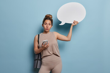 Photo of impressed sporty woman with hair bun dressed in brown tracksuit holds mobile phone and blank speech bubble for your advertising content carries rolled karemat isolated over blue background