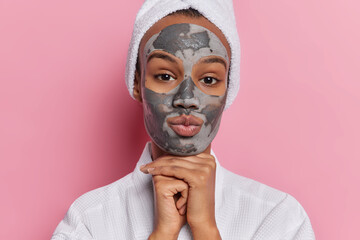 Beauty and spa procedures. Studio shot of young glad European lady standing isolated in centre on pink background keeping hands under chin applying grey natural clay face mask to reduce wrinkles