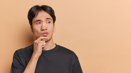 People emotions concept. Young thoughtful Asian man holding left hand on chin trying to solve difficult task or problem standing on left isolated on beige background with blank space for advertisement