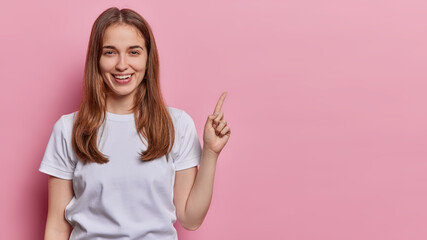 Horizontal shot of pleased cheerful woman with straight hair points index finger on blank space dressed in casual white t shirt shows advetisement isolated ove pink background. Look at this.