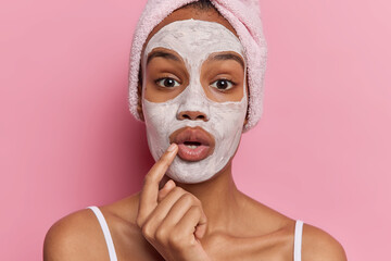 Photo of surprised young female model holds breath looks stunned applies facial beauty mask for skin treatment wears bath towel on head stands bare shouldered isolated over pink studio background