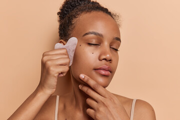 Photo of curly haired African girl does guasha face massage with jade stone enjoys spa procedures at home keeps eyes closed wears t shirt isolated over brown background. Facial care concept.