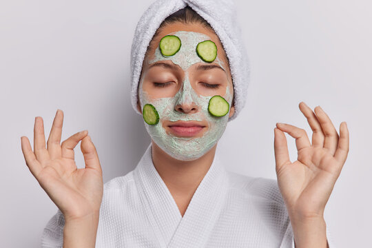 Horizontal shot of calm young woman immerses herself in moment of rejuvenation applies facial clay mask with cucumbers keeps eyes closed wears bathrobe and towel on head isolated over white background - Powered by Adobe
