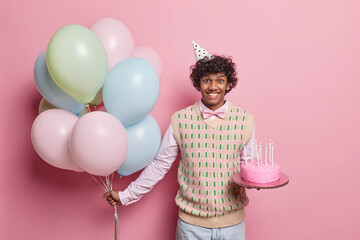 Cheerful birthday Hindu man holds bunch of vibrant helium balloons in one hand and sweet treat...
