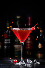 Manhattan alcoholic cocktail drink with bourbon, red vemuth, bitter, ice and cocktail cherry in glass, dark bar counter background, bar tools and bottles