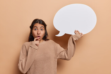 Horizontal shot of impressed Iranian girl with dark hair holding empty speech bubble for your advertising content has mouth opened from surprisement wears knitted jumper isolated over brown background
