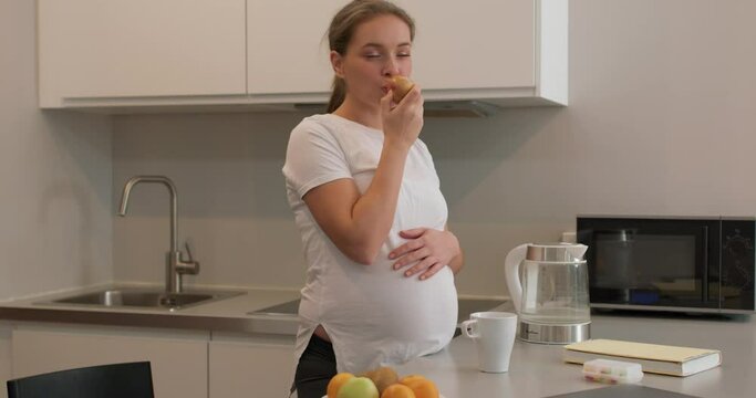 Pregnant woman standing in the kitchen and eating an apple, An apple a day keeps a doctor away Slow motion health care, wellness