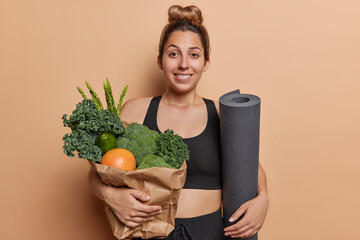 Horizontal shot of sporty young woman with hair bun bites lips carries paper bag full of fresh...