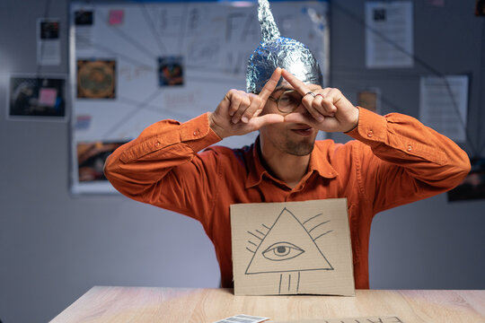 Young stupid man in cap of aluminum foil holding banner with all-sees eye. Concept of a conspiracy theory. Conspiracy.