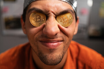 Strange stupid man conspiracy theorist in protective foil cap holds two bitcoins in his eyes. Male with bitcoins in eyes. Bitcoin is a cryptocurrency. Fake news concept - obrazy, fototapety, plakaty