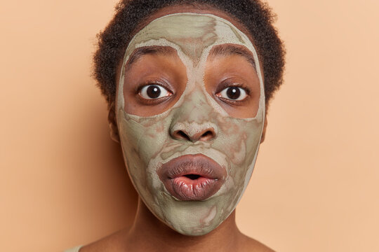Beauty and cosmetology concept. Studio close up of young surprised African female seeing something unusual or unexpected looking straight at camera applying clay face mask to reduce wrinkles