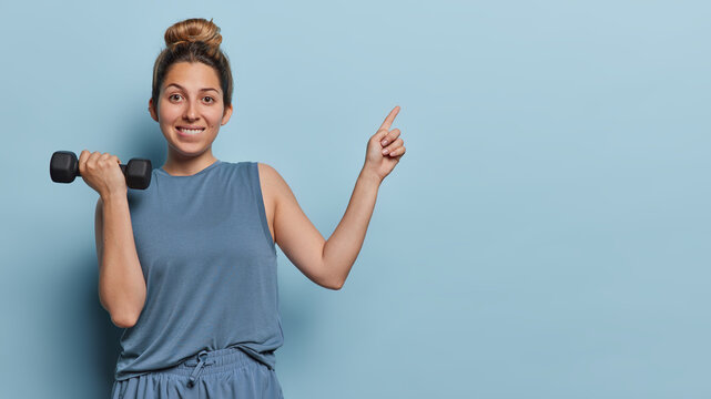 Horizontal shot of sporty young woman raises dumbbell does physical exercises points index finger on copy space for your advertising content dressed in sportswear isolated over blue background.