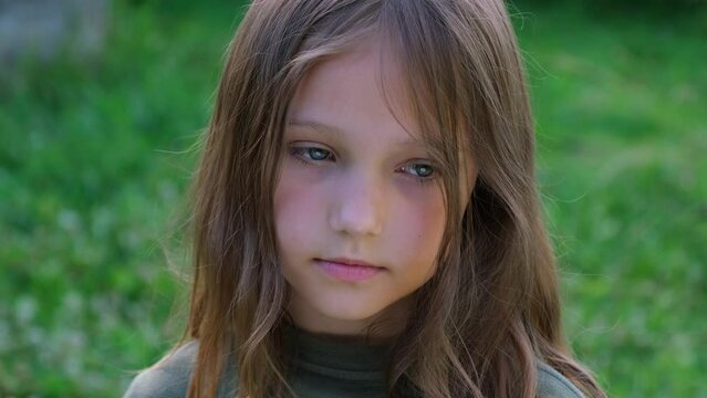 Portrait of a little beautiful girl looking at the camera, standing on the street in the city on a summer day. Young sad thinking curious child looking at camera closeup outdoors. Face Eyes Serious