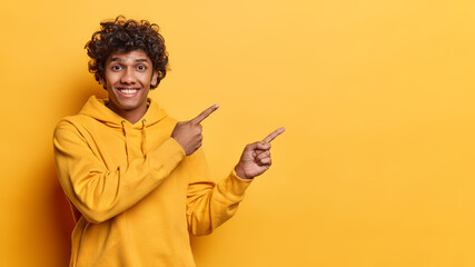 Positive emotions concept. Studio waist up of young cheerful smiling broadly Hindu guy wearing hoodie standing on left isolated on yellow background pointing at blank space for your advertisement - 626482108