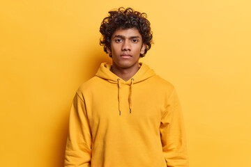 People and positive emotions concept. Studio photo of young calm glad peaceful Hindu male with curly hair standing isolated in centre on yellow background wearing casual clothes keeping hands down © WHstudio Leushin N