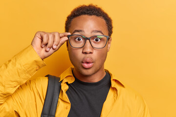 Portrait of handsome curly haired teenage man keeps hand on rim of spectacles keeps lips rounded...