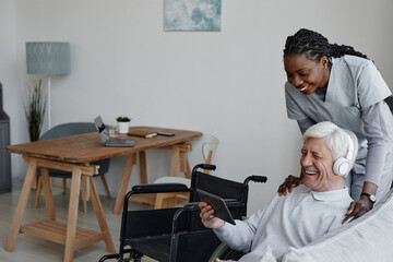 Portrait of smiling senior man with disability having fun at home and using tablet with nurse assisting, copy space
