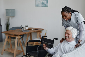 Portrait of smiling senior man with disability having fun at home and using tablet with nurse...