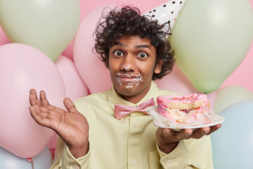 Puzzled clueless Hindu man raises palm with hesitation poses with cake has mouth smeared with cream...
